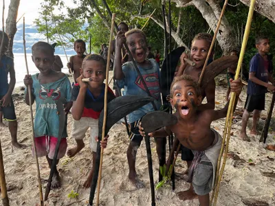 Children holding wooden paddles and poles gather beneath a beachside tree.