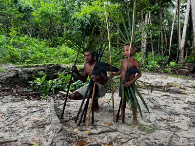 Two children stand among palms with poles and leaf decorations.