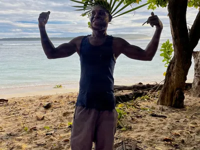 A man wearing a leaf headdress raises his arms beside the sea.