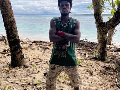 A man stands with folded arms beneath a beachside tree.