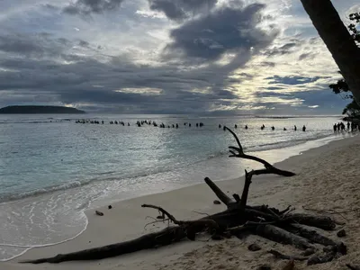 People gather in shallow water beneath a cloudy morning sky.