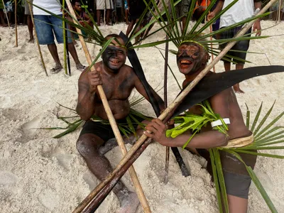 Two participants wear leaf headdresses and hold poles on the beach.