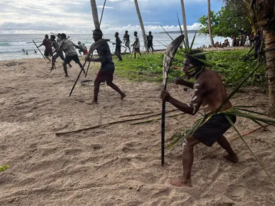 Participants face each other with long poles on the beach.