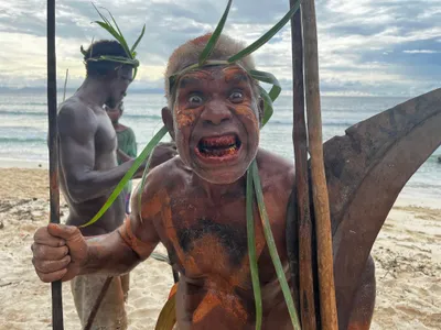 A participant with painted face and leaf headdress smiles at the camera.