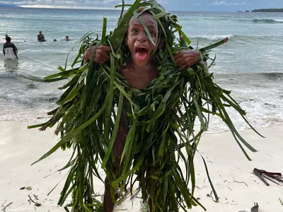 A participant covered in green leaves poses beside the water.