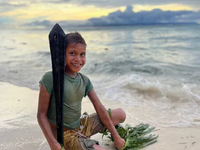 A smiling child holds a wooden paddle at the shoreline.