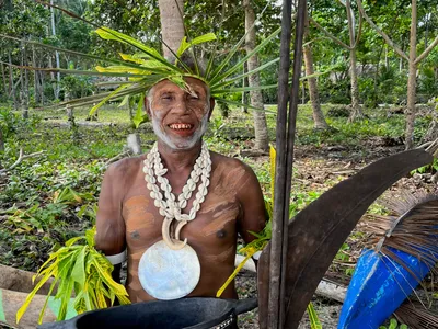 A smiling participant wears a shell necklace and leaf headdress.