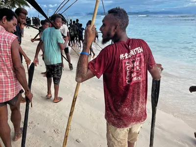 A line of participants carrying poles follows the shoreline.
