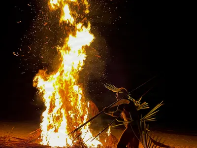 A tall bonfire lights participants and their poles at night.