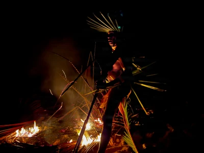 A participant wearing leaf decorations stands beside a bonfire.