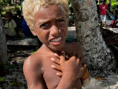 A child with painted face stands beside a tree.