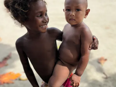 A child holds a younger child beside the beach.